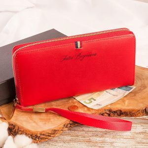 Czerwony portfel z zamkiem błyskawicznym, z logo 'Jestem Bezcenna', leżący na drewnianym podłożu obok kilku banknotów i monety.