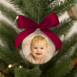 Szklana bombka świąteczna z uśmiechniętą twarzą niemowlęcia w beżowym sweterku, zawieszona na choince, z czerwoną kokardą i napisem 'Nikola' w złotym kolorze.