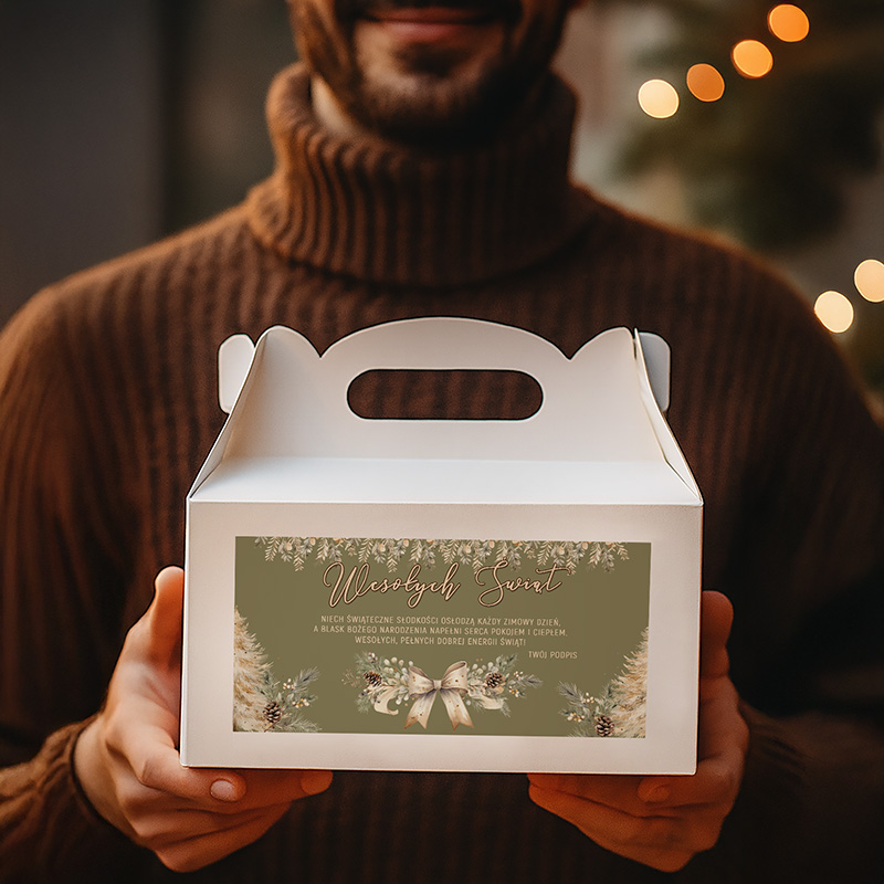 Mężczyzna z brązowym swetrem trzyma biały kartonowy pojemnik z uchwytem, na którym znajduje się dekoracyjny projekt z napisem 'Wesołych Świąt' oraz motywami bożonarodzeniowymi, w tle widoczne delikatne światełka choinkowe.
