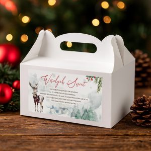 Pudelko-swiateczne-na-prezent-z-grafika-jelenia-i-zimowym-lasem Biała pudełko z uchwytem, na którym widnieje tekst "Wesołych Świąt" oraz dekoracja z reniferem i świątecznymi roślinami, umieszczone na drewnianym blacie obok szyszek i choinkowych ozdób w tle.