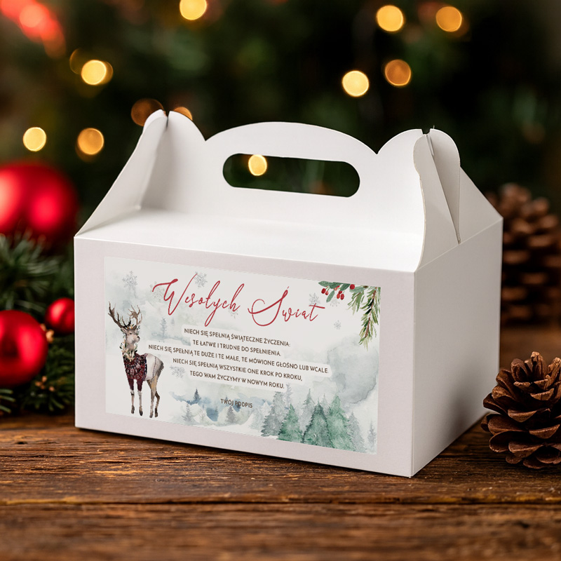 Biała pudełko z uchwytem, na którym widnieje tekst "Wesołych Świąt" oraz dekoracja z reniferem i świątecznymi roślinami, umieszczone na drewnianym blacie obok szyszek i choinkowych ozdób w tle.