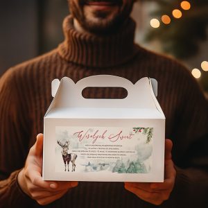 Mężczyzna w brązowym swetrze trzyma białe pudełko z uchwytem, na którym znajduje się świąteczny motyw z reniferem i napisem 'Wesołych Świąt'. W tle widać świąteczne iluminacje.