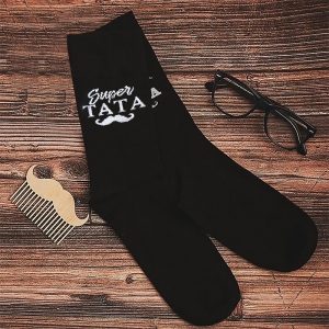 Czarne skarpetki z napisem 'Super Tata' i muszką, obok drewniany grzebień w kształcie wąsów oraz okulary na drewnianym tle.