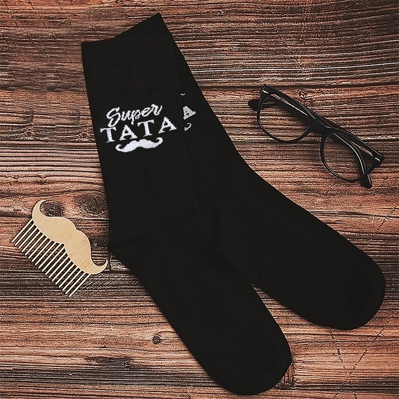 Czarne skarpetki z napisem 'Super Tata' i muszką, obok drewniany grzebień w kształcie wąsów oraz okulary na drewnianym tle.