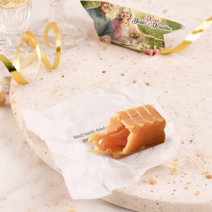 Mały kawałek cukierków na jasnym, marmurowym talerzu, otoczony dekoracyjną złotą wstążką; w tle widoczna kartka z napisem 'Dzień Babci i Dziadka' i wizerunkiem uśmiechniętej pary, a pod cukierkiem fragment papieru z napisanym cytatem w języku polskim.