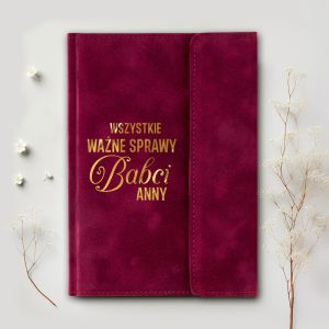 Fioletowy notes z napisem "WSZYSTKIE WAŻNE SPRAWY Babci ANNY" na okładce, otoczony delikatnymi, białymi kwiatkami i gałązkami na jasnym tle.
