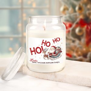 Świeca zapachowa w słoiku z etykietą przedstawiającą Świętego Mikołaja w saniach ciągniętych przez renifera, z napisem 'HO HO HO' oraz tekstem 'Zapal i oficjalnie rozpocznij Święta'. Świeca płonie, a tło jest rozmyte z choinką i połyskującymi ozdobami.