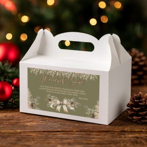 swiateczne-pudelko-na-ciasto-z-grafika-i-zyczeniami Biała pudełko na prezenty z napisem 'Wesołych Świąt' oraz dekoracyjnym motywem bożonarodzeniowym, umieszczone na drewnianym stole z ozdobami w tle, w tym choinkowym gałązkami i bombkami.
