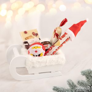 Biała saneczka wypełniona słodkościami, w tym czekoladą z owocami, słoikiem Nutelli, i zabawką w kształcie Mikołaja, z tłem w rozmytych białych i złotych światełkach.