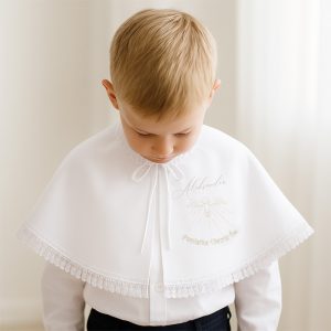 Chłopiec z blond włosami noszący białą pelerynkę z haftem i kokardką, patrzy w dół, otaczają go jasne tło i zasłony.