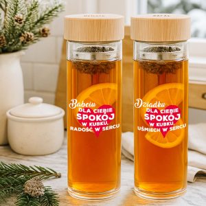 Dwa przezroczyste kubki z herbatą, z widokiem na plaster cytryny w każdej szklance. Na jednym kubku napis 'Babciu dla Ciebie spokój w kubku, radość w sercu', a na drugim 'Dziadku dla Ciebie spokój w kubku, uśmiech w sercu'. W tle rośliny i jasne otoczenie wnętrza.