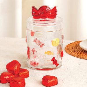 Słój szklany z czerwonym deklem w kształcie cukierka, ozdobiony kolorowymi cukierkami, stojący na jasnej powierzchni, obok leżą kilka czerwonych cukierków.