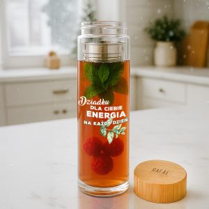 Przezroczysty bidon szklany na napój zawierający ciepły napój z liśćmi mięty i malinami, z napisem 'Dziadku dla Ciebie energia na każdy dzień'. W tle kuchnia z jasnymi elementami.