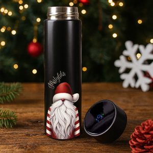 Czarny termos z ilustracją zatkanego Santy z długą białą brodą i czerwonym kapeluszem, z napisem 'Matylda', leżący na drewnianym stole obok czarnego pokrywki z cyfrowym wyświetlaczem; w tle dekoracje bożonarodzeniowe, w tym zielone gałązki i świąteczne światła.