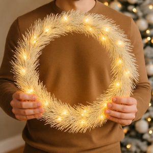Mężczyzna w brązowej bluzie trzyma ozdobny wieniec z jasnymi lampkami, na tle choinki z białymi i srebrnymi dekoracjami.
