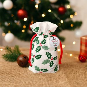 Mała torba ozdobna z motywem świątecznym, pokryta zielonymi liśćmi ostrokrzewu i czerwoną jagodą, z czerwonymi sznurkami u góry oraz okrągłą etykietą z napisem 'Wesołych Świąt!', na tle choinki z kolorowymi światłami i ozdobami.