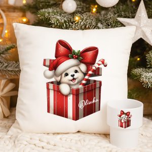 Poduszka z ilustracją szczeniaka w czerwonym pudełku prezentowym z białą kokardą i czapką Mikołaja, trzymającego laskę cukrową; obok filiżanka z podobnym wzorem; w tle choinka z ozdobami.