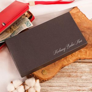 Czarna, elegancka paczka z napisem "Kochanej Babci Kasi" leży na drewnianym stoliku obok czerwonego portfela z wystającym banknotem 50 złotych i monetą. W tle widać biały materiał i bawełnę.