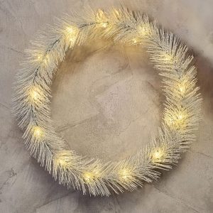 Okrągły wieniec dekoracyjny z białych igiełek, subtelnie podświetlony ciepłymi białymi światłami, umieszczony na lekko teksturowanej, jasnej powierzchni.