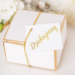 Biała pudełko z złotymi akcentami, ozdobione złotą nitką, z białą kartką z napisem 'Dziękujemy'. W tle widoczne kwiaty.