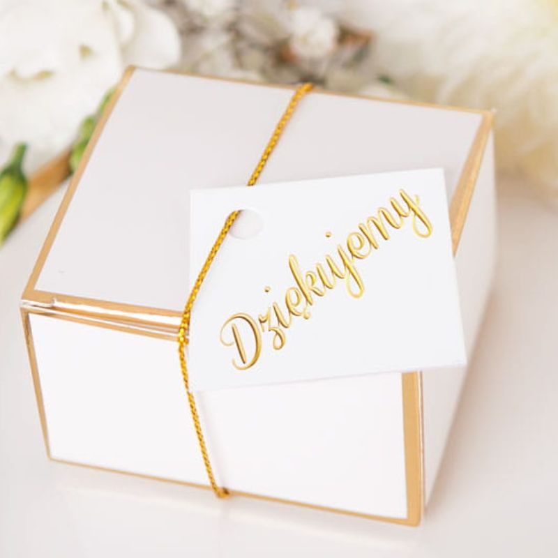 Biała pudełko z złotymi akcentami, ozdobione złotą nitką, z białą kartką z napisem 'Dziękujemy'. W tle widoczne kwiaty.