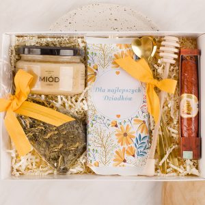 Zestaw prezentowy z miodem, suszem ziołowym, kartką z napisem 'Dla najlepszych Dziadków', łyżką i miodologią zestawionymi w eleganckim opakowaniu z żółtą wstążką.