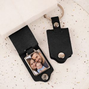 Czarny brelok do kluczy z metalowym uchwytem, w którym umieszczone jest zdjęcie rodziny składającej się z matki, ojca i dziecka.