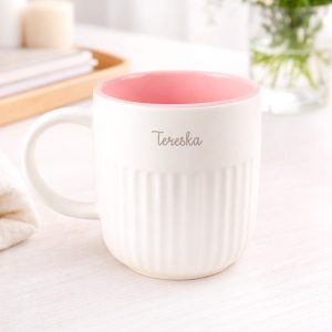 ceramiczny-kubek-dla-babci-z-rozowym-wnetrzem Biała filiżanka z różowym wnętrzem, z wyszczerbionymi krawędziami i napisem 'Terenska' w jasnym kolorze, stoi na drewnianym blacie obok ręcznika i bukietu kwiatów w wazonie.