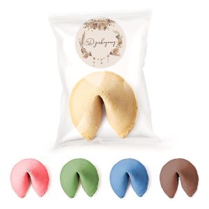 Opakowanie z ciastkiem fortuny w kolorze beżowym znajduje się na przezroczystym tle, obok niego leżą cztery ciastka w różnych kolorach: różowym, zielonym, niebieskim i brązowym. Na opakowaniu widnieje okrągła etykieta z napisem 'Dziękujemy' oraz ozdobami roślinnymi.