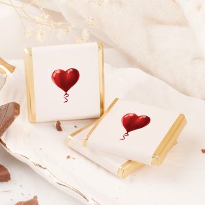 Dwa pakunki słodyczy o białym opakowaniu ozdobionym czerwonym balonem w kształcie serca, leżące na eleganckim talerzu, w otoczeniu drobnych dekoracji i czekolady.