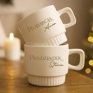 Dwie białe filiżanki ustawione jedna na drugiej, z napisem 'Prababcia Aria' na górnej filiżance oraz 'Pradziadek Stasiu' na dolnej, w tle delikatne oświetlenie i świeczka.