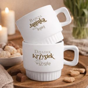 Dwie białe filiżanki na drewnianym talerzu, z napisami: "Babcia Janinka wie lepiej" na górnej filiżance i "Dziadek Krzyśiek wie wszystko" na dolnej, otoczone słodkościami i lampkami świec w tle.