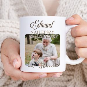 Kubek trzymany w dłoniach, na którym widnieje napis "Edmund NAJLEPSZY PRADZIADek na świecie" oraz zdjęcie przedstawiające starszego mężczyznę z siwymi włosami i małego chłopca, którzy siedzą na trawie.