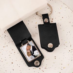 fotobrelok-na-prezent-dla-pary.jpg Czarny breloczek z miejscem na zdjęcie, w którym znajduje się zdjęcie pary całującej się na tle białej sukni i bukietu kwiatów. Brelok leży na jasno nakrapianym tle.