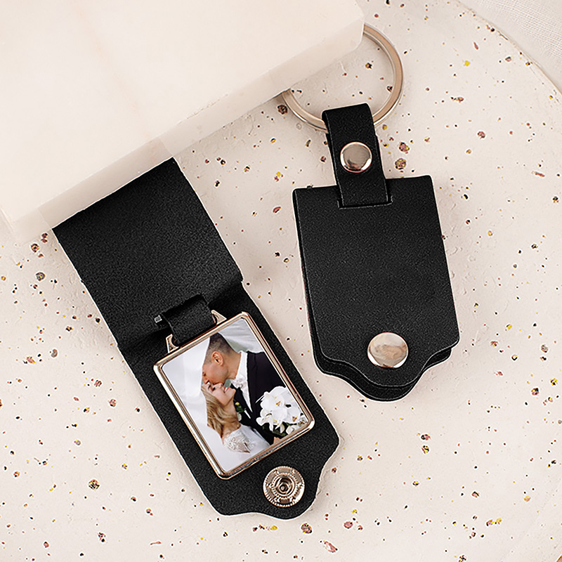 Czarny breloczek z miejscem na zdjęcie, w którym znajduje się zdjęcie pary całującej się na tle białej sukni i bukietu kwiatów. Brelok leży na jasno nakrapianym tle.