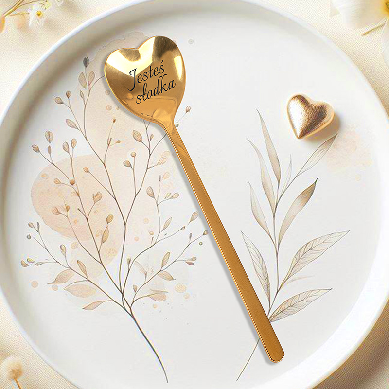 Złota łyżka w kształcie serca z wygrawerowanym napisem 'Jesteś słodka', umieszczona na talerzu z delikatnym wzorem roślinnym w jasnych kolorach. Obok łyżki znajduje się małe złote serce.