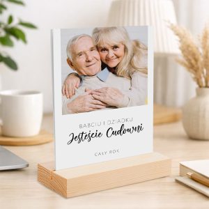 Zdjęcie przedstawiające starszą parę, uśmiechających się do aparatu, przytulających się na tle neutralnego tła. Obok nich widnieje napis "Babciu i Dziadku Jesteście Cudowni" oraz "Cały Rok". Całość umieszczona jest na drewnianym stojaku w przytulnym wnętrzu.