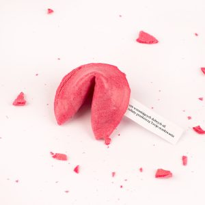Różowy ciasteczko fortuny, częściowo otwarte, z przesłaniem widocznym na białym kawałku papieru, otoczone okruchami na białym tle.