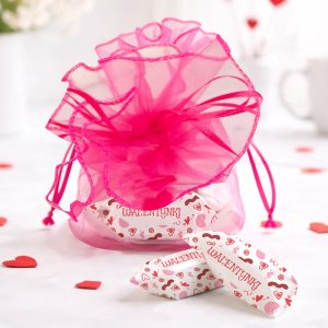 Różowa torebka prezentowa z ozdobnym, siateczkowym wykończeniem i zawiązanym sznurkiem, w której znajdują się cukierki z napisem 'WALENTYNKI', leżące na jasnym tle z serduszkami w różnych kolorach.