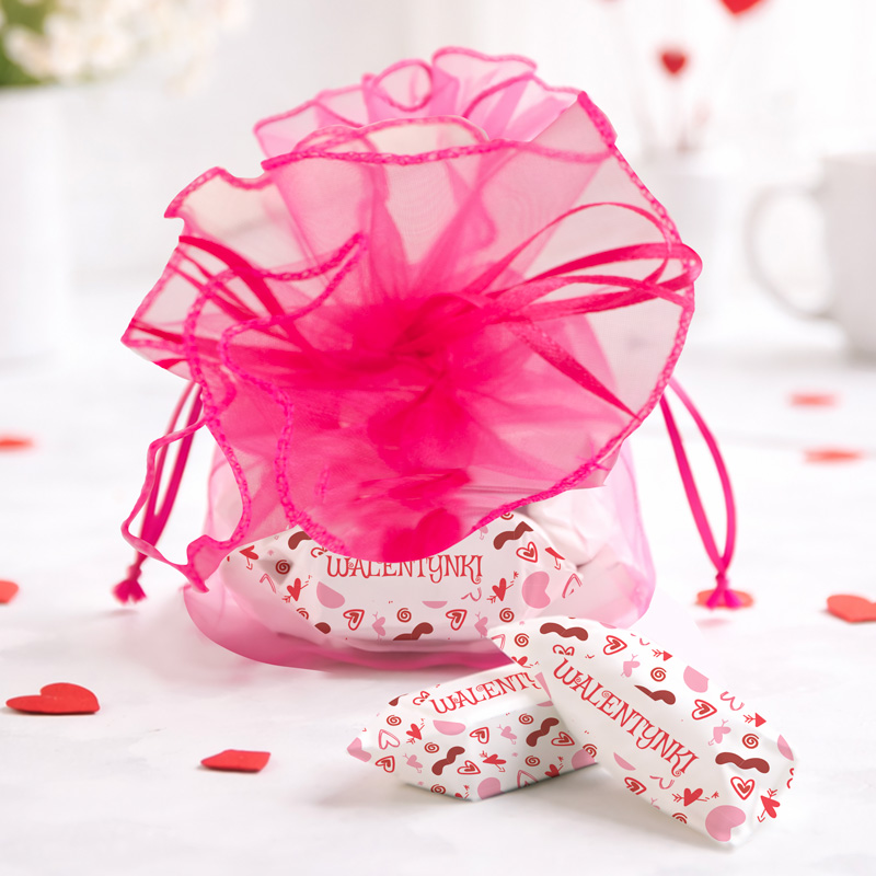 Różowa torebka prezentowa z ozdobnym, siateczkowym wykończeniem i zawiązanym sznurkiem, w której znajdują się cukierki z napisem 'WALENTYNKI', leżące na jasnym tle z serduszkami w różnych kolorach.