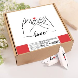 Pudełko z grafiką przedstawiającą splecione dłonie i czerwone serce, z napisem 'love' na froncie, stoi na jasnym tle, obok znajduje się mała, biała karteczka z tym samym napisem.