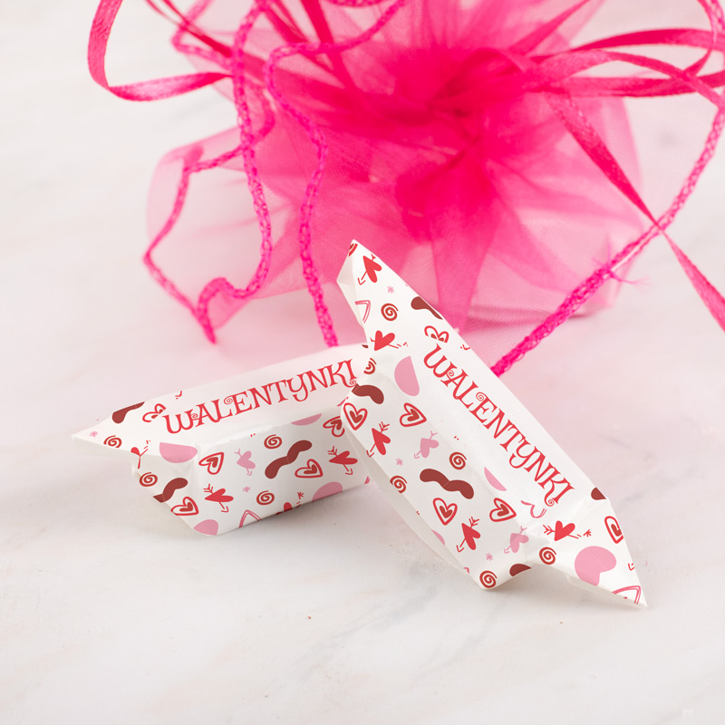 Dwa białe kartoniki z napisami 'WALENTYNKI', pokryte kolorowymi wzorami serc, ust i innych symboli miłości, leżą na jasnym tle obok różowej wstążki z organzy.