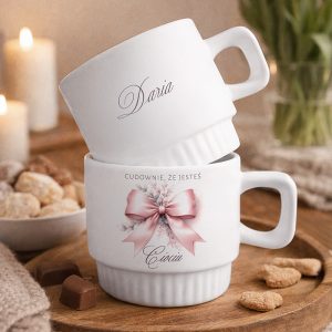 Dwie białe filiżanki ustawione jedna na drugiej, z napisem "Daria" na górnej filiżance i napisem "CUDOWNIE, ŻE JESTEŚ Ciociu" na dolnej, otoczone słodkościami i świece w tle.