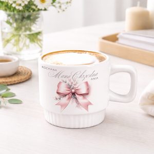 Biały kubek z napisem "Kochana Mama Chrzestna Ania" oraz dekoracyjną różową wstążką, z kremowym napojem na wierzchu, stojący na jasnym stole w tle z kwiatami i świeczką.
