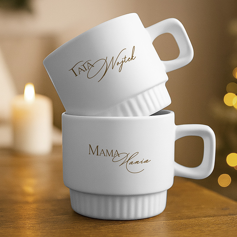 Dwie białe filiżanki ustawione jedna na drugiej, z napisem 'Tata Wojtek' na górnej filiżance i 'Mama Hania' na dolnej, na drewnianym blacie, w tle delikatne światło świecy.