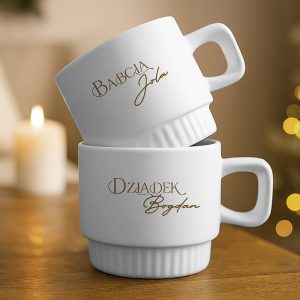 Dwa białe kubki z brązowymi napisami na froncie, górny kubek ma napis 'Babcia Jola', a dolny 'Dziadek Bogdan', ustawione na drewnianym stole, w tle rozmyte światła i świeca.