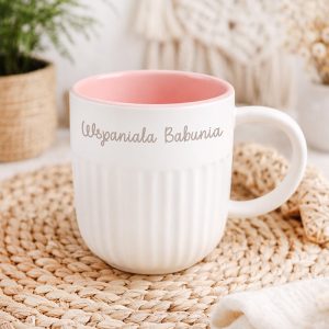 Biały kubek z różowym wnętrzem, z tekstem "Wspaniała Babunia" na powierzchni, umieszczony na podkładce z plecionki, w tle widoczna roślinność oraz dekoracje domowe.