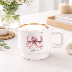 Biały kubek z różową wstążką i napisem "CUDOWNIE, ŻE JESTEŚ". Kubek stoi na jasnym stole obok filiżanki kawy, owinięty w słoneczny, minimalistyczny styl z kwiatową dekoracją w tle.