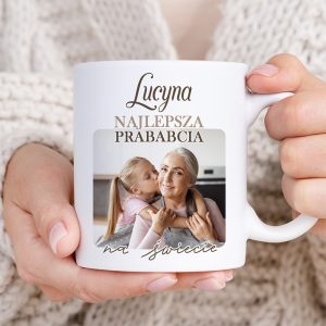 Kobieta trzyma filiżankę z napisem "Lucyna Najlepsza Prababcia na świecie", zbliżenie na twarz uśmiechniętej starszej kobiety i małej dziewczynki, która ją całuje; w tle kuchnia.