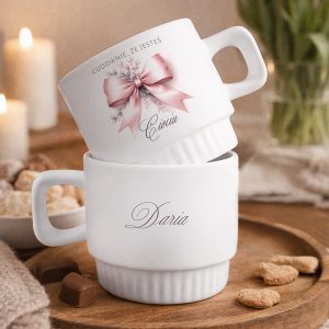 Dwie białe filiżanki ustawione jedna na drugiej, z ozdobnymi napisami i różową kokardą, na tle świec i słodkości.